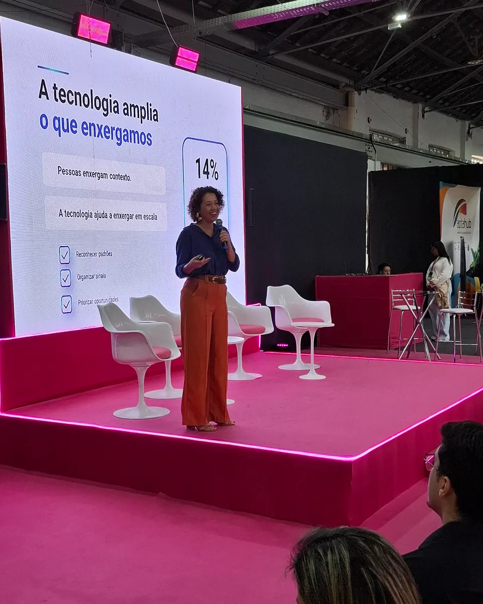 Pessoa com microfone no palco, diante de uma projeção e de pessoas na plateia.